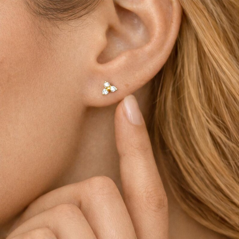 May include: A close-up of a gold stud earring with three small, white and yellow gemstones. The earring is on a person's ear. The person's finger is touching their ear.