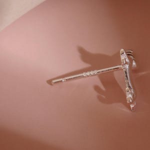 Op de afbeelding: Zilverkleurige oorbel met een uniek ontwerp, met een rechte pin met kleine decoratieve accenten en een gedetailleerde klauwachtige vorm aan het uiteinde. De oorbel is geplaatst tegen een zachtroze achtergrond.