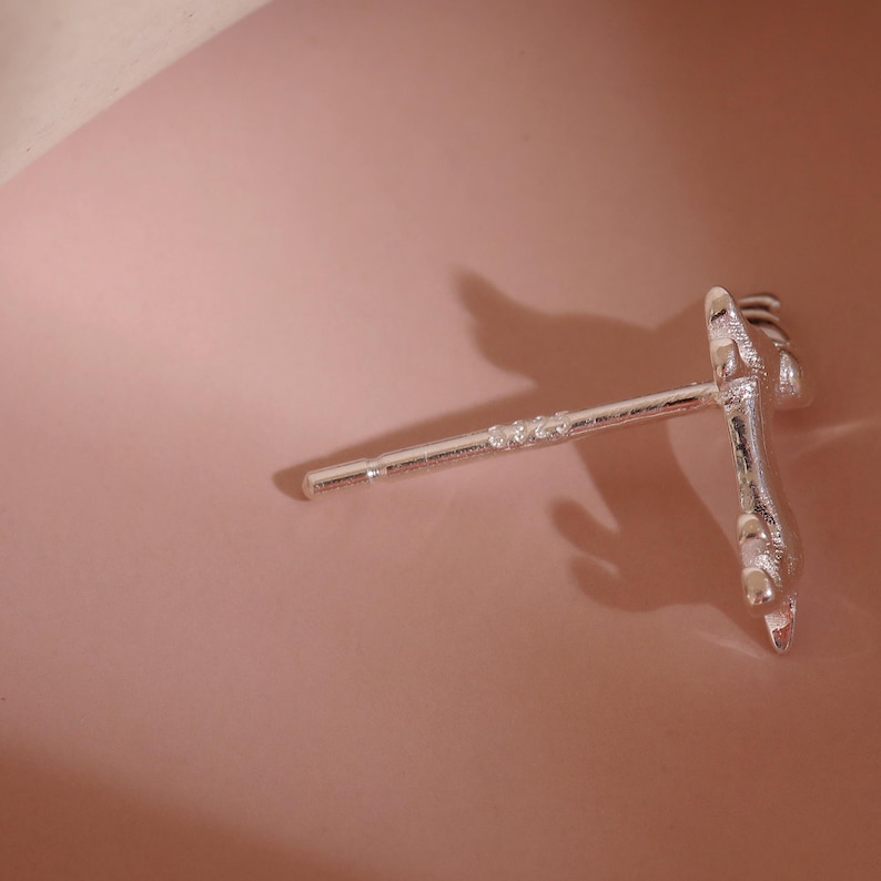 Op de afbeelding: Zilverkleurige oorbel met een uniek ontwerp, met een rechte pin met kleine decoratieve accenten en een gedetailleerde klauwachtige vorm aan het uiteinde. De oorbel is geplaatst tegen een zachtroze achtergrond.