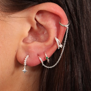 May include: A close-up of a person's ear with multiple silver earrings. The earrings include a small hoop with a green stone, a larger hoop with a chain attached, and a smaller hoop with a star charm.