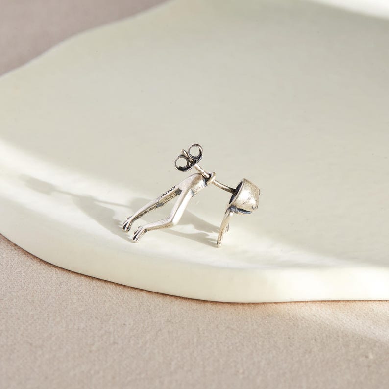 May include: Silver earring featuring a stylized human figure in a crouching pose. The earring is designed to be worn on the earlobe.