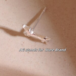Op de afbeelding: Een zilveren oorbel met een abstract ontwerp. De oorbelpen is zichtbaar. De tekst "AG stands for Store Brand" staat onderaan de afbeelding. De oorbel ligt op een lichtbruin oppervlak.