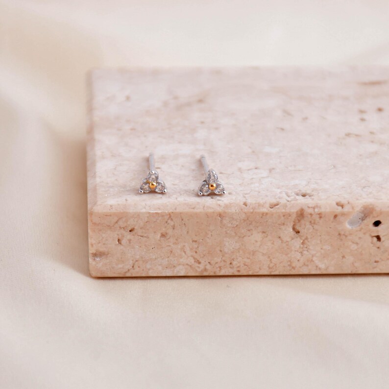 May include: A pair of small stud earrings. Each earring features a silver post with a gold-colored center and three clear, round accent stones arranged in a triangular shape. The earrings are displayed on a light-colored, textured surface.