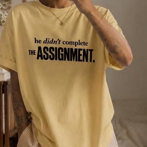 May include: A pale yellow short-sleeved t-shirt featuring the black printed text 'he didn't complete THE ASSIGNMENT.' The shirt has a crew neckline and a relaxed fit, paired with light-coloured vertical striped trousers with a subtle texture.