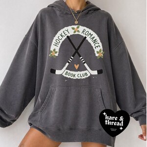 Op de afbeelding: Donkergrijze hoodie met een voorzak. De afbeelding toont "Hockey Romance Book Club" met gekruiste hockeysticks, bloemmotieven en een hart. Het merklogo "hare & thread" staat rechtsonder.