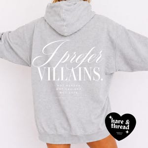 May include: A light gray hoodie with the text "I prefer VILLAINS" in white script. Below the text are the words "NOT HEROES, NOT SAVIORS, NOT SAFE." A black heart-shaped logo with the words "hare & thread" is in the lower right corner.