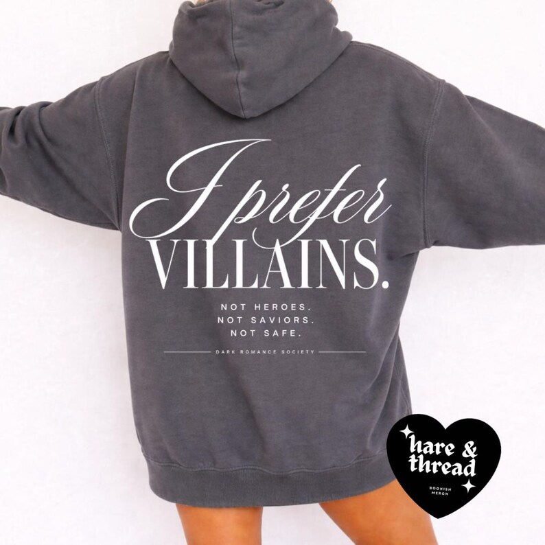May include: A dark gray hoodie with the text "I Prefer Villains" in white script. Below, it reads "Not Heroes. Not Saviors. Not Safe." The hoodie features a drawstring hood and a kangaroo pocket.