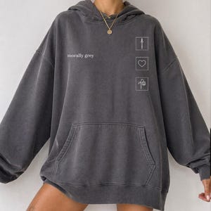 May include: Dark grey hoodie with a front pocket and a hood. The text "morally grey" is printed on the front, along with three small white graphic icons: a knife, a heart, and a flower. The hoodie has a relaxed fit.