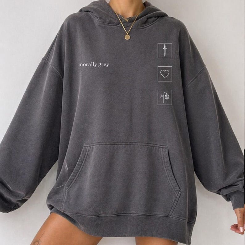 May include: Dark grey hoodie with a front pocket and a hood. The text "morally grey" is printed on the front, along with three small white graphic icons: a knife, a heart, and a flower. The hoodie has a relaxed fit.