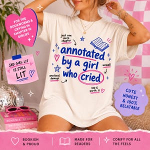 Op de afbeelding: Cr&egrave;mekleurig T-shirt met de tekst "annotated by a girl who cried" in blauw en roze. Het shirt heeft extra tekst en afbeeldingen, waaronder harten, sterren en de woorden "Sad Girl Lit is Still Lit".
