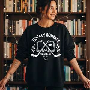 May include: Black sweatshirt with the white text "HOCKEY ROMANCE BOOK CLUB EST 2025". The design includes crossed hockey sticks, an open book with a heart, and laurel branches. The sweatshirt is worn by a person.