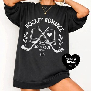 Op de afbeelding: Zwart sweatshirt met de witte tekst "HOCKEY ROMANCE BOOK CLUB EST 2025". Het ontwerp bevat gekruiste hockeysticks, een open boek met een hart en een hockeypuck. Een zwart hartvormig logo met de tekst "hare & thread" staat rechtsonder.