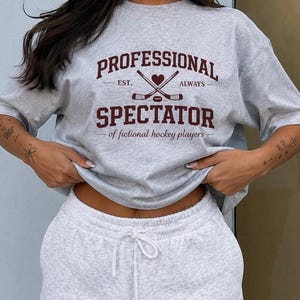 Puede incluir: Una camiseta de cuello redondo gris claro con texto en rojo oscuro que dice PROFESSIONAL SPECTATOR, junto a un gráfico de dos palos de hockey cruzados, un corazón y un disco. Debajo, el texto dice of fictional hockey players.