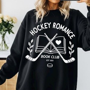 May include: Black sweatshirt with the text "HOCKEY ROMANCE BOOK CLUB EST 2025" in white. The design includes crossed hockey sticks, a heart, an open book, and a hockey puck. The sweatshirt is worn by a person wearing a pink beanie and sunglasses.