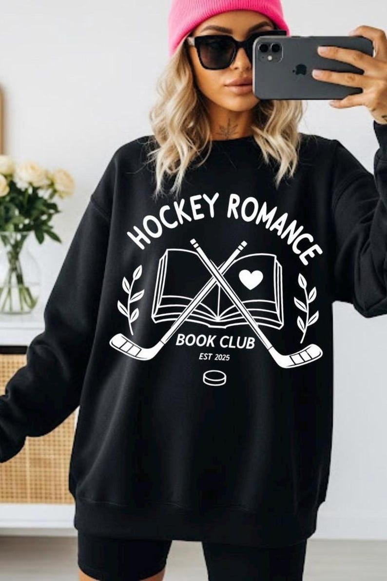 May include: Black sweatshirt with the text "HOCKEY ROMANCE BOOK CLUB EST 2025" in white. The design includes crossed hockey sticks, a heart, an open book, and a hockey puck. The sweatshirt is worn by a person wearing a pink beanie and sunglasses.