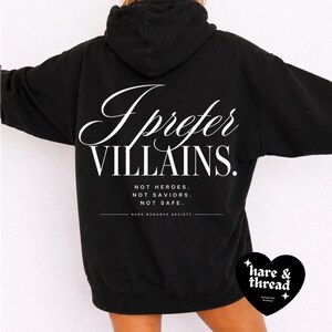 May include: Black hoodie with the text "I Prefer Villains" in white script. Below the main text are the words "NOT HEROES. NOT SAVIORS. NOT SAFE." The hoodie has a drawstring hood and a kangaroo pocket. The brand logo is in the bottom right corner.