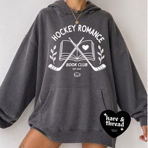 Op de afbeelding: Donkergrijze hoodie met een voorzak en een witte opdruk met de tekst "HOCKEY ROMANCE BOOK CLUB EST 2025" met gekruiste hockeysticks, een boek en een hart. Het merklogo "hare & thread" staat rechtsonder.