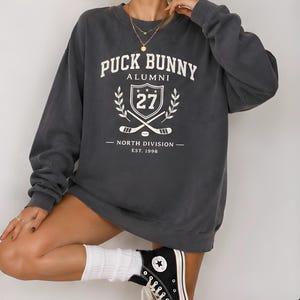 Op de afbeelding: Een antracietgrijze sweater met ronde hals en witte opdruk met de tekst PUCK BUNNY ALUMNI, NORTH DIVISION en EST. 1998. De afbeelding bevat een schild met het getal 27, gekruiste hockeysticks en accenten van laurierbladeren.