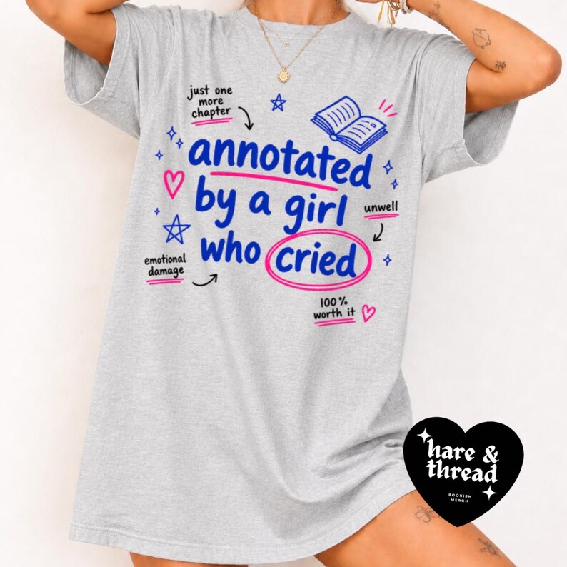 Op de afbeelding: Lichtgrijs t-shirt met de tekst "annotated by a girl who cried" in blauw en roze. Het ontwerp bevat illustraties van een boek, sterren en harten. De tekst "100% worth it" is ook aanwezig.