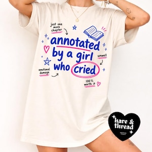 Op de afbeelding: Cr&egrave;mekleurig T-shirt met de tekst "annotated by a girl who cried" in blauw en roze, omringd door krabbels en de woorden "just one more chapter" en "100% worth it". Onderaan staat een zwart hartvormig logo.