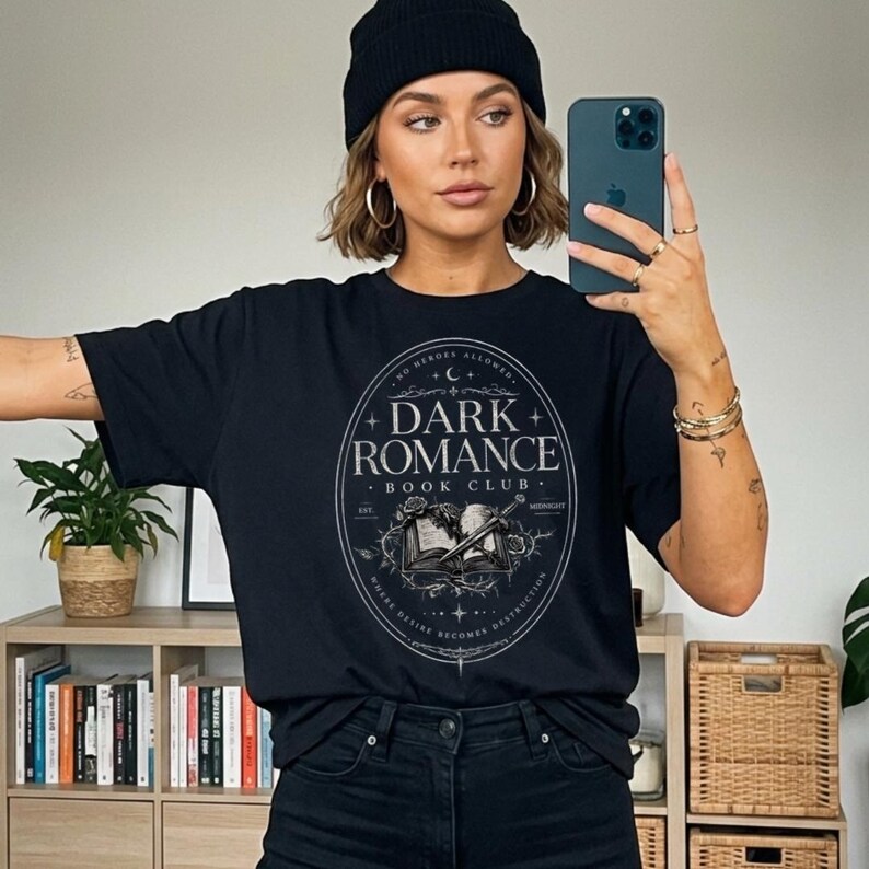 Puede incluir: Camiseta negra con un dise&ntilde;o ovalado blanco y gris que dice "DARK ROMANCE BOOK CLUB" con una ilustraci&oacute;n de un libro y una espada. La camiseta tambi&eacute;n tiene el texto "NO HEROES ALLOWED" y "WHERE DESIRE BECOMES DESTRUCTION".