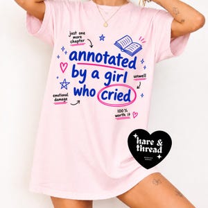 Op de afbeelding: Lichtroze t-shirt met de tekst "annotated by a girl who cried" in blauw en roze. Het ontwerp bevat een boekillustratie en de woorden "emotional damage" en "unwell".