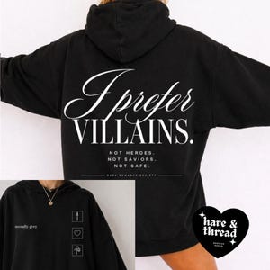 May include: Black hoodie with the text "I prefer VILLAINS." in white script on the back. Below the text are the words "NOT HEROES. NOT SAVIORS. NOT SAFE." A smaller image shows the hoodie in grey with the text "morally grey".