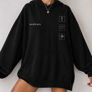 May include: Black hoodie with a front pocket and a drawstring hood. The text "morally grey" is printed on the front. Three white line drawings of a knife, heart, and flower are on the right side of the hoodie.