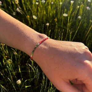 Könnte beinhalten: Ein Perlenarmband mit roten, grünen und weißen Perlen. Das Armband wird an einem Handgelenk getragen, mit einem Hintergrund aus grünem Gras und weißen Blumen.