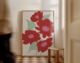 May include: Large framed art print featuring stylized red flowers with pink centers and green leaves against a cream background. The artwork is in a gold frame and leans against a white wall. A small wooden side table is next to the artwork.