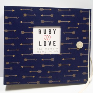 May include: Navy blue baby book with a gold arrow pattern. The cover has a square cutout with the words "RUBY LOVE" and "the modern baby book". A cream-colored button and ribbon closure are on the right side.