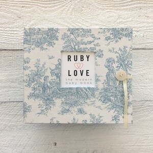May include: A white baby book with a blue toile pattern and a white frame with the text "Ruby Love the modern baby book".