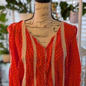 May include: A red blouse with a white dotted pattern and a V-neck. The blouse has long sleeves and is trimmed with cream-colored lace along the front and sleeves. It is buttoned down the front and has a gathered waist.