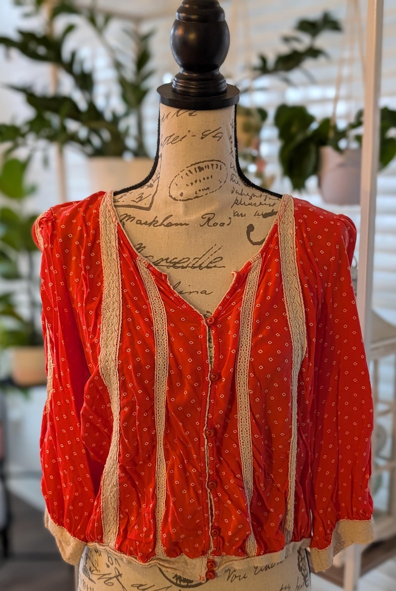 May include: A red blouse with a white dotted pattern and a V-neck. The blouse has long sleeves and is trimmed with cream-colored lace along the front and sleeves. It is buttoned down the front and has a gathered waist.