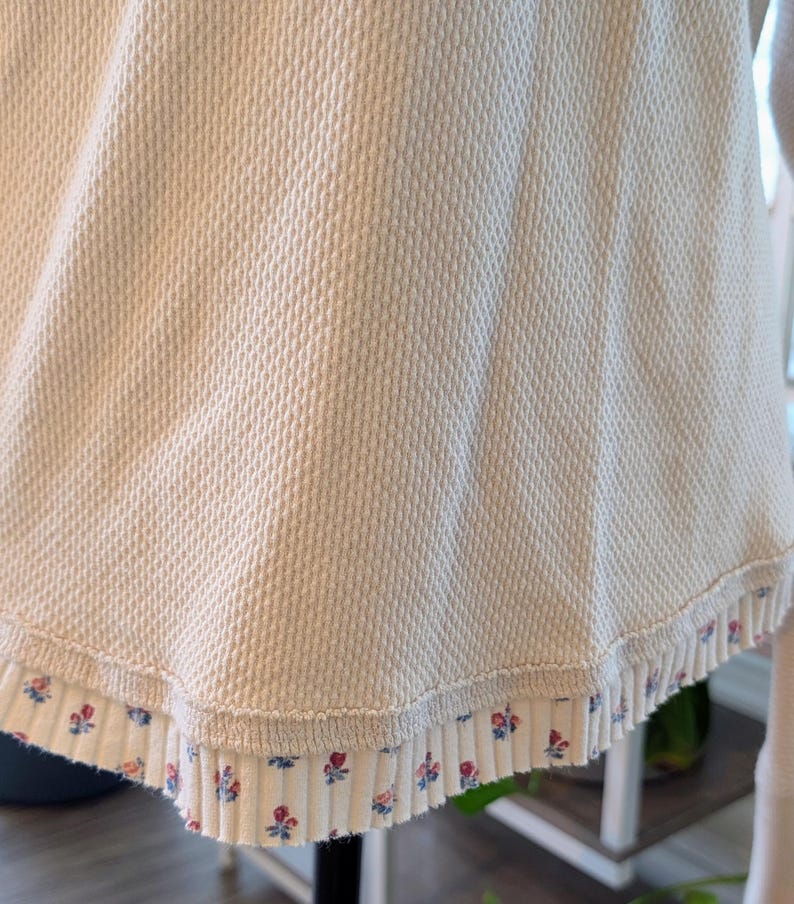 Puede incluir: Primer plano de una parte superior de punto de gofre color crema con un ribete floral. El ribete presenta peque&ntilde;as flores rojas y azules sobre un fondo blanco. La tela tiene un patr&oacute;n texturizado en forma de rejilla.