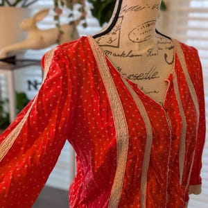 May include: A red blouse with a white dotted pattern and cream-colored lace trim. The long-sleeved top has a V-neck and is gathered at the waist. The blouse is displayed on a mannequin.