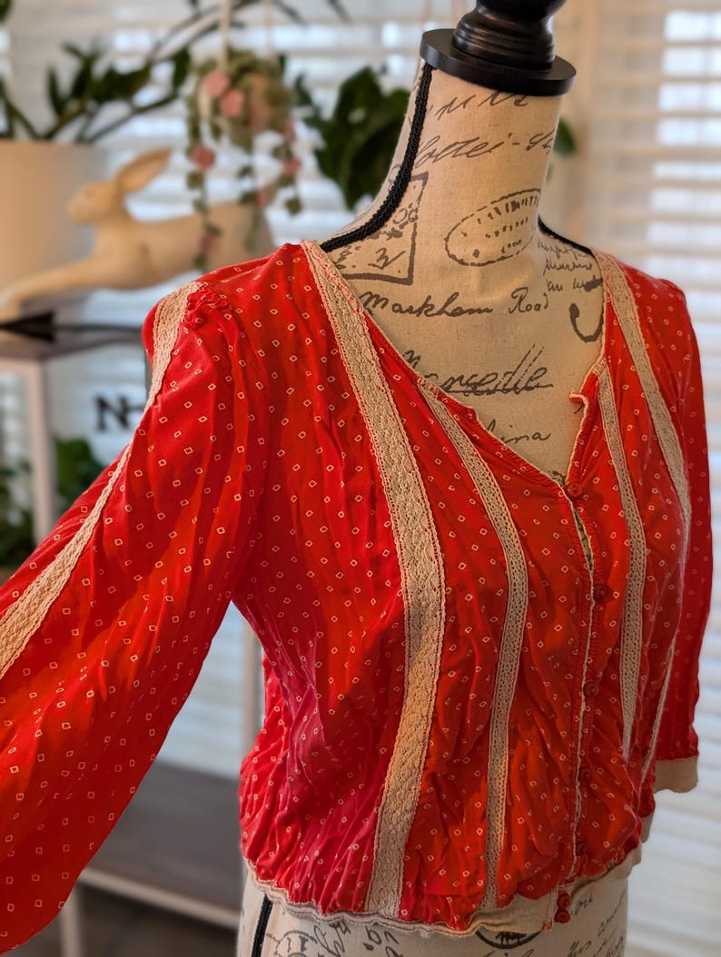 May include: A red blouse with a white dotted pattern and cream-colored lace trim. The long-sleeved top has a V-neck and is gathered at the waist. The blouse is displayed on a mannequin.