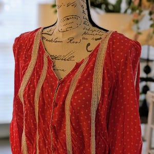 May include: A red button-down blouse with a white polka dot pattern. The blouse features cream-colored lace trim along the front and sleeves. The garment is displayed on a mannequin.