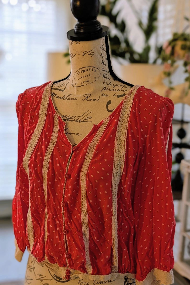 May include: A red button-down blouse with a white polka dot pattern. The blouse features cream-colored lace trim along the front and sleeves. The garment is displayed on a mannequin.