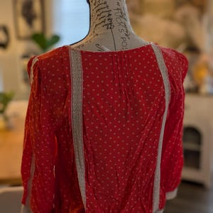 May include: Red blouse with white polka dots and white lace trim. The long-sleeved top is displayed on a mannequin. The blouse has a gathered waist and a round neckline. The sleeves end with a white trim.