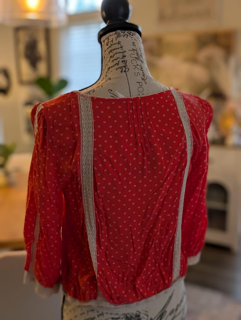 May include: Red blouse with white polka dots and white lace trim. The long-sleeved top is displayed on a mannequin. The blouse has a gathered waist and a round neckline. The sleeves end with a white trim.