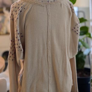 Puede incluir: Camisa beige de manga larga de punto de gofre con detalles florales en las mangas y el escote. La camisa tiene un corte holgado y un dobladillo curvo. La parte trasera de la camisa presenta un detalle de costura vertical.