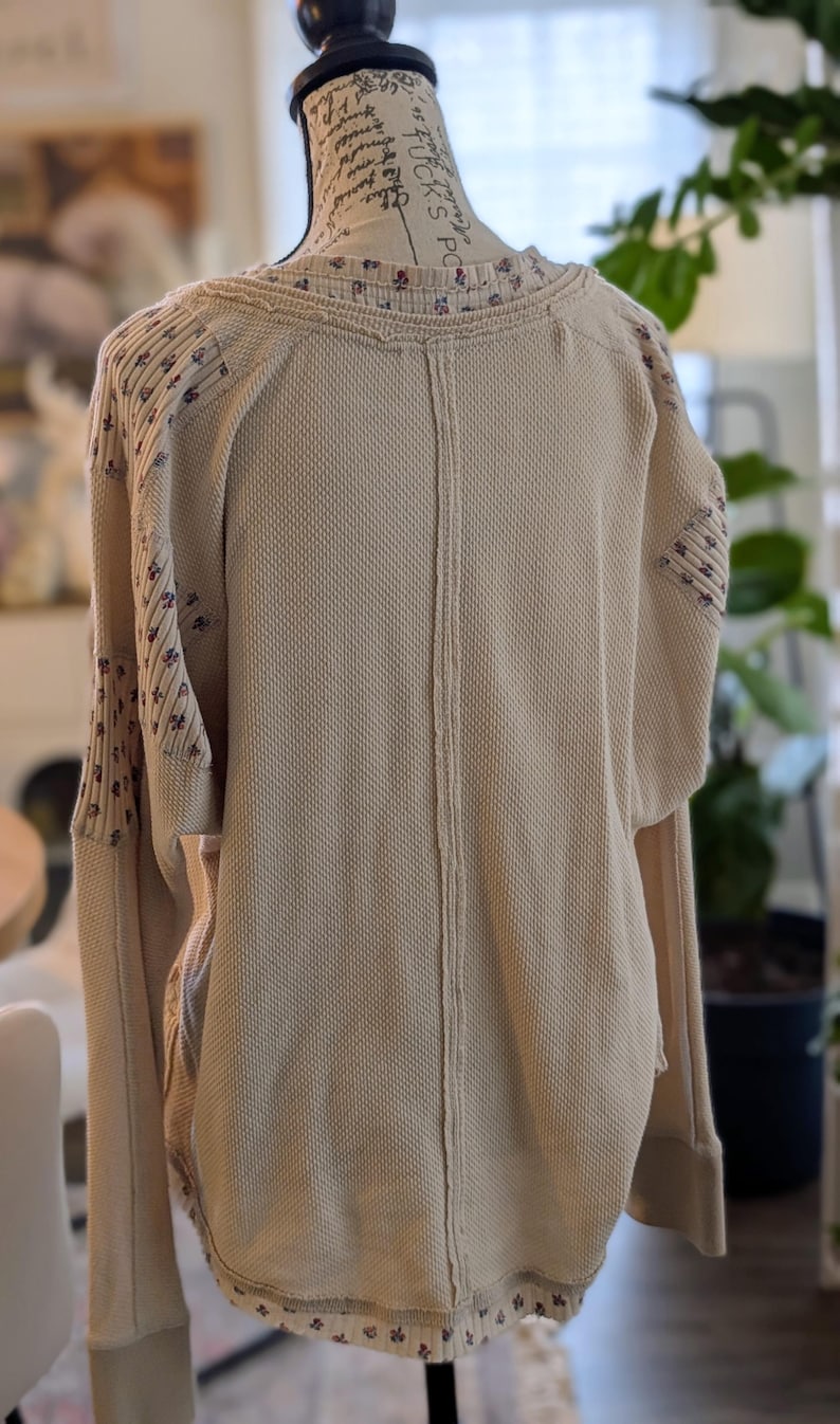 Puede incluir: Camisa beige de manga larga de punto de gofre con detalles florales en las mangas y el escote. La camisa tiene un corte holgado y un dobladillo curvo. La parte trasera de la camisa presenta un detalle de costura vertical.