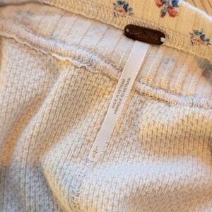 Puede incluir: Primer plano de una tela acanalada de color crema con un estampado floral de flores azules y rojas. Se adjunta una etiqueta marr&oacute;n con las palabras "Free People". Tambi&eacute;n se adjunta una etiqueta blanca con texto.