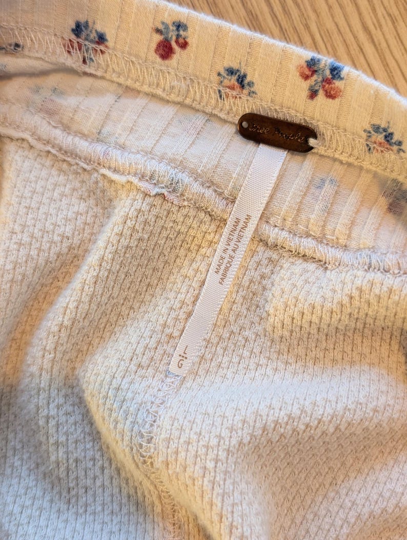 Puede incluir: Primer plano de una tela acanalada de color crema con un estampado floral de flores azules y rojas. Se adjunta una etiqueta marr&oacute;n con las palabras "Free People". Tambi&eacute;n se adjunta una etiqueta blanca con texto.