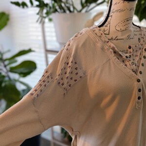 Puede incluir: Camisa henley de punto de gofre color crema con parches estampados florales en las mangas y el escote. La camisa tiene una parte delantera abotonada y un corte holgado. La tela tiene un aspecto texturizado, y el estilo general es informal y c&oacute;modo.