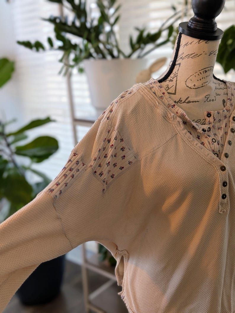 Puede incluir: Camisa henley de punto de gofre color crema con parches estampados florales en las mangas y el escote. La camisa tiene una parte delantera abotonada y un corte holgado. La tela tiene un aspecto texturizado, y el estilo general es informal y c&oacute;modo.