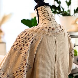 Puede incluir: Una blusa beige de punto de gofre con mangas y cuello estampados florales se exhibe en un maniqu&iacute;. Las mangas presentan un dise&ntilde;o de patchwork con peque&ntilde;os detalles florales rojos y azules. La blusa tiene un aspecto texturizado y un corte holgado.