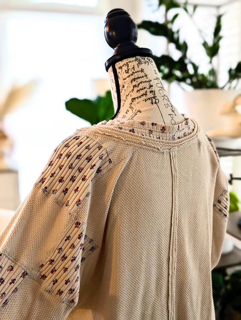 Puede incluir: Una blusa beige de punto de gofre con mangas y cuello estampados florales se exhibe en un maniqu&iacute;. Las mangas presentan un dise&ntilde;o de patchwork con peque&ntilde;os detalles florales rojos y azules. La blusa tiene un aspecto texturizado y un corte holgado.