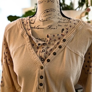 Puede incluir: Una camisa henley beige de punto de gofre con una capa interior y mangas con estampado floral. La camisa tiene una parte delantera abotonada y mangas largas. La cabeza del maniqu&iacute; es visible en el fondo.